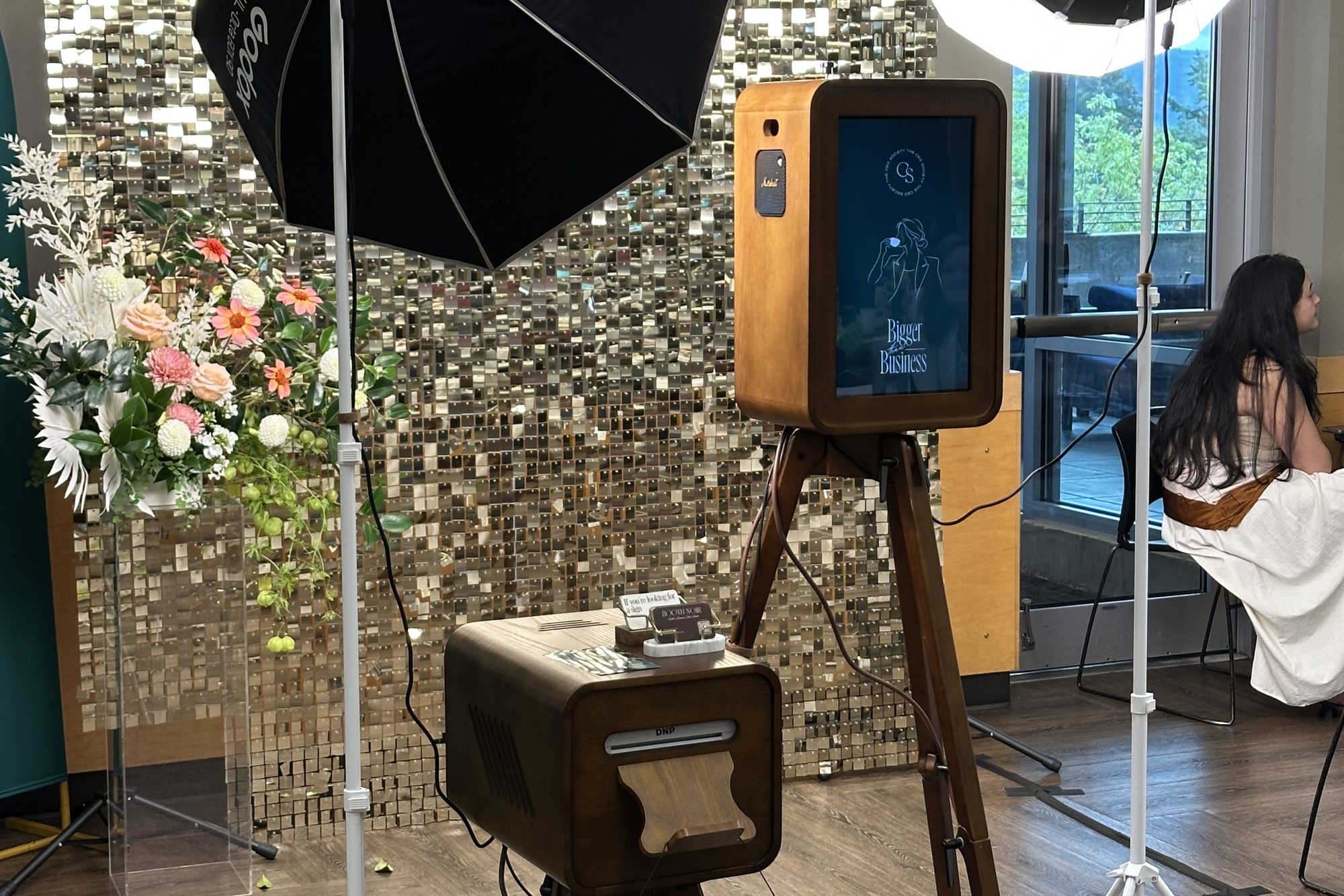 The stained-wood booth on its tripod in a venue, lit by a softbox and a ring light, with flowers and a sequin wall behind